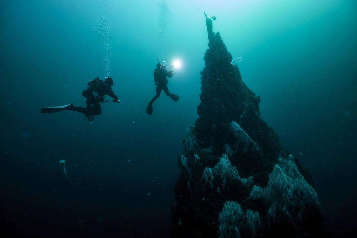 Divers with a light beside a submerged pinnacle in PADI Deep Diver imagery. © PADI 2026