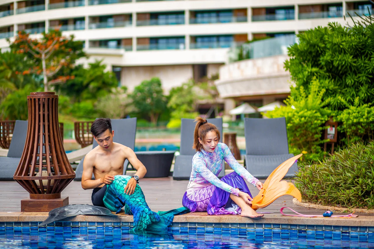 Two people in mermaid tails sit beside a pool in PADI Mermaid referral-program imagery.